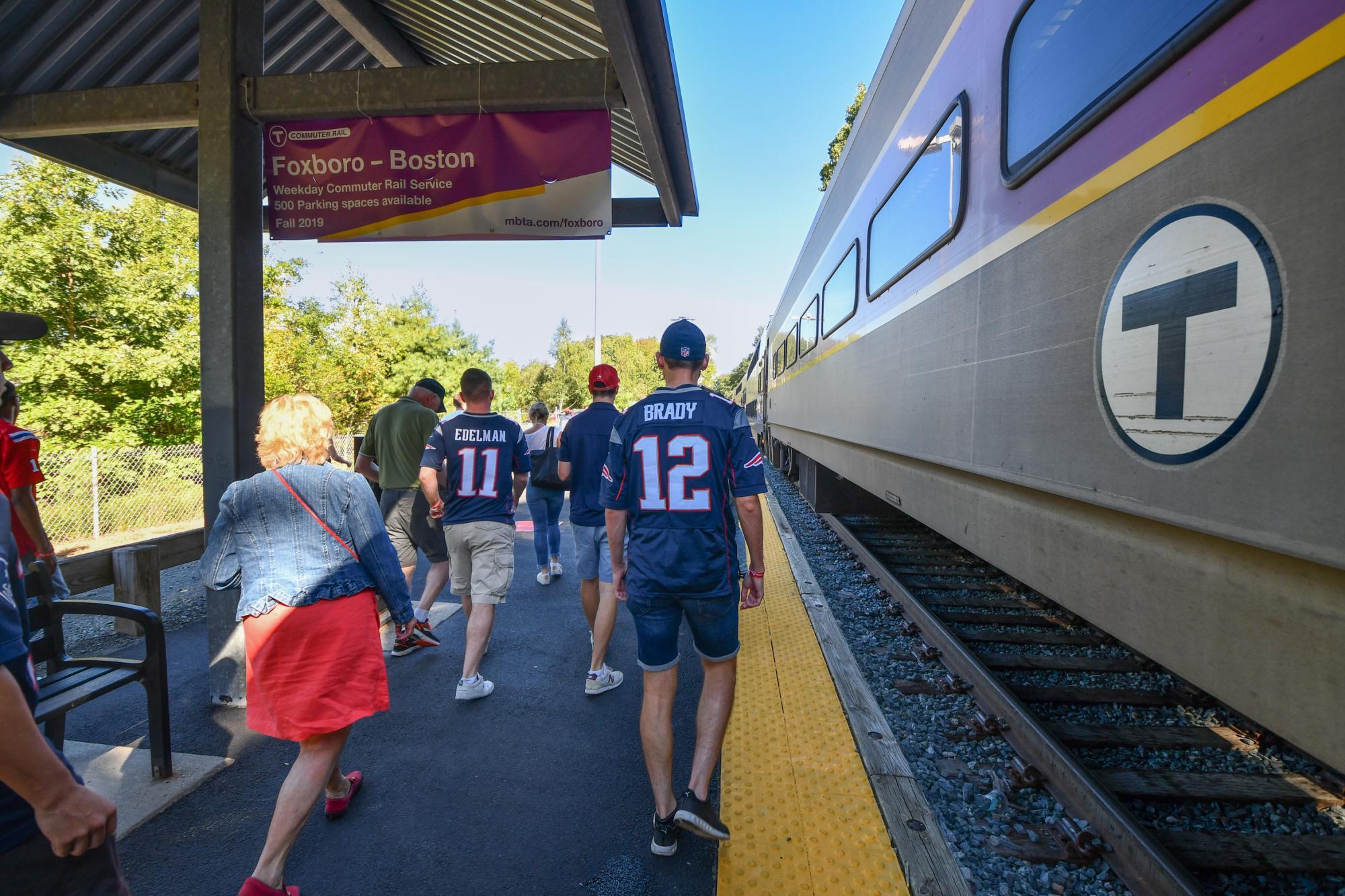 At Foxboro Station, riders with Patriots jerseys walk on the platform, passing by the Commuter Rail train.