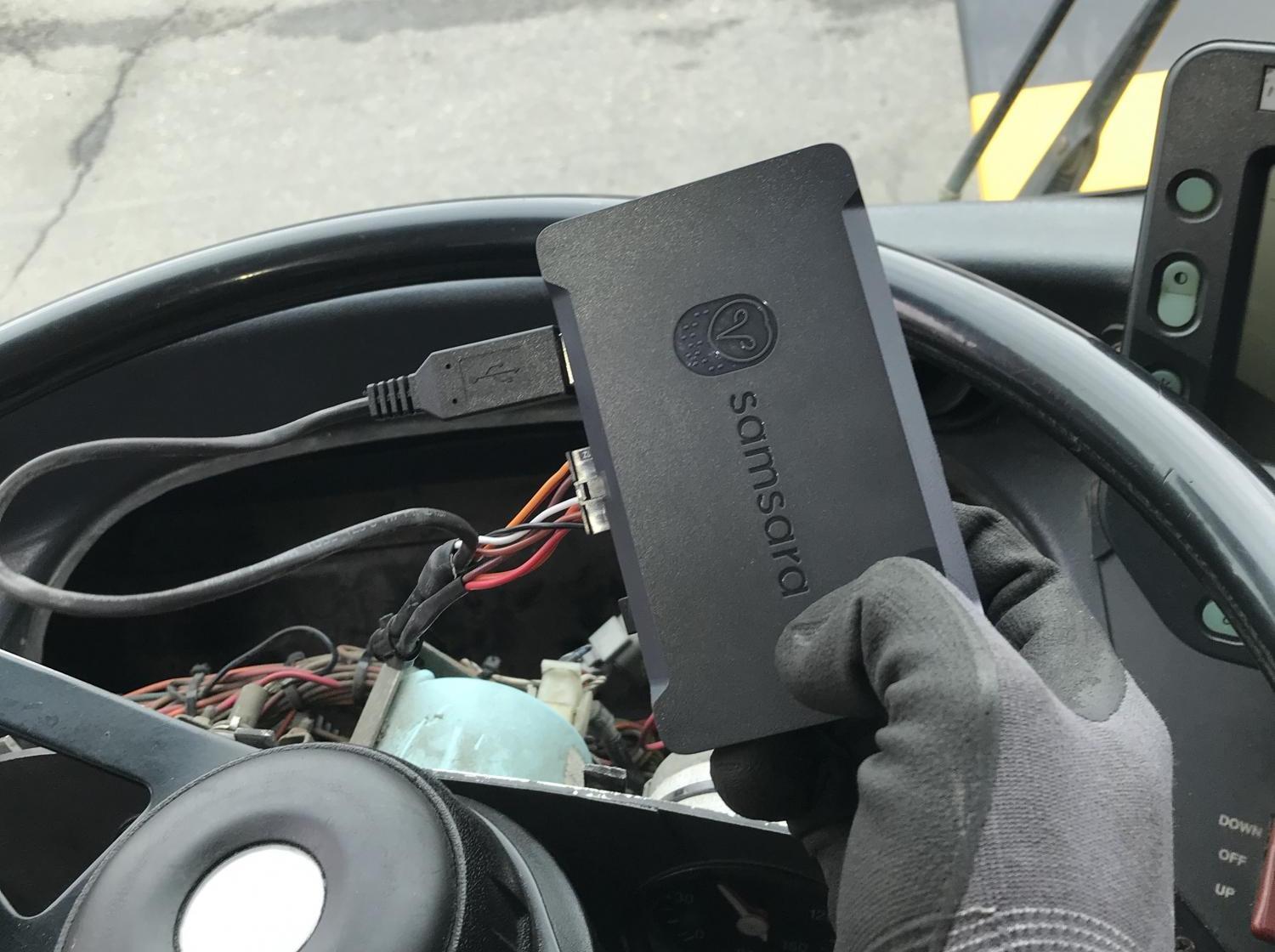 Closeup of a Samsara GPS device, held by a gloved driver's hand in front of a bus steering wheel.
