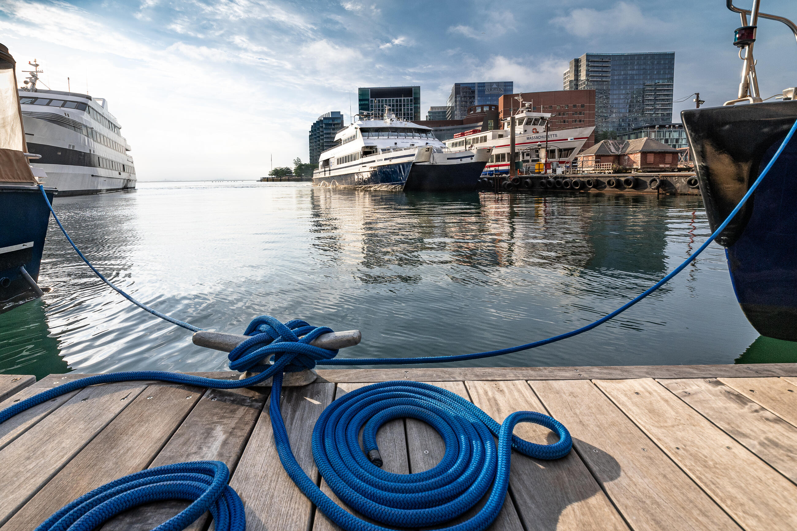 A photo of a ferry in the water, taken from the Rowes Wharf dock. There's a large blue rope tied to the dock in the foreground.
