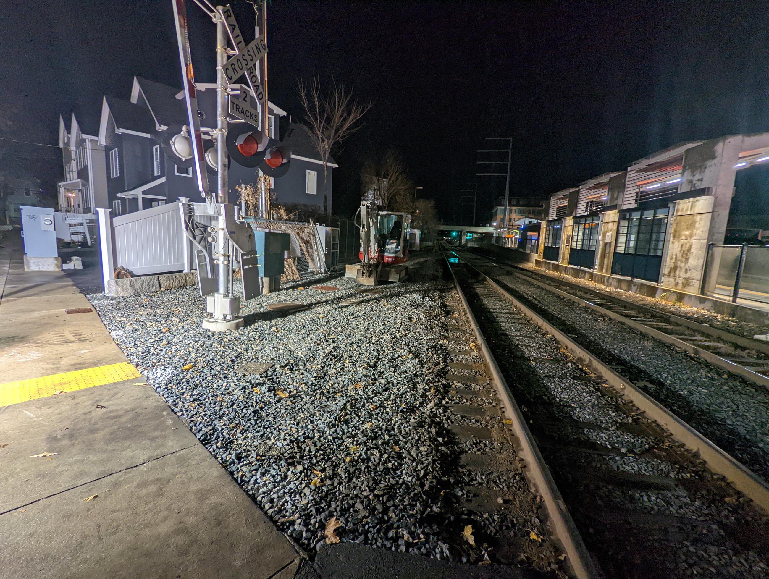 The site of the old Chelsea station is shown in a photo taken at night. At left is the railroad crossing with a fence and houses behind it, and a sidewalk to its left. In front of the crossing is a patch of gravel. At right is a train track. At far right is the back of the new Chelsea station. 