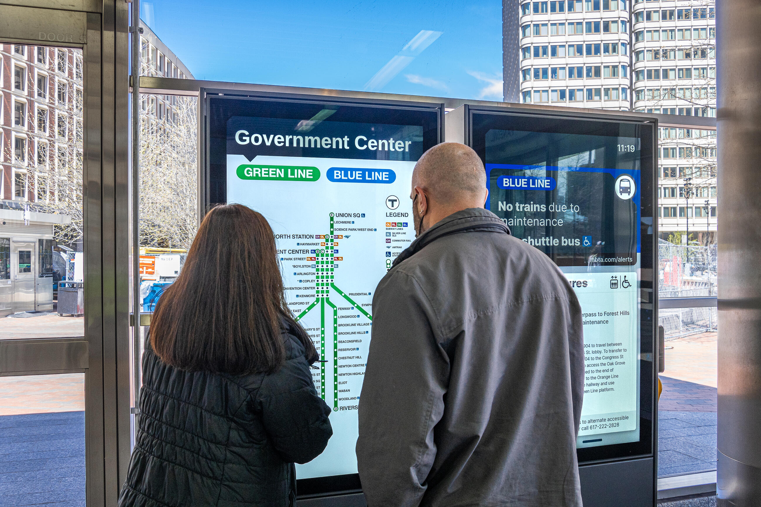 Two riders at Government Center face a two-screen digital sign. At left is a screen showing the Green Line map with a large button that allows users to switch to a Blue Line map. At right is a screen that shows a Blue Line alert saying no trains are running and that shuttles are running instead. Below that is a second alert about other Blue Line closures due to maintenance. 