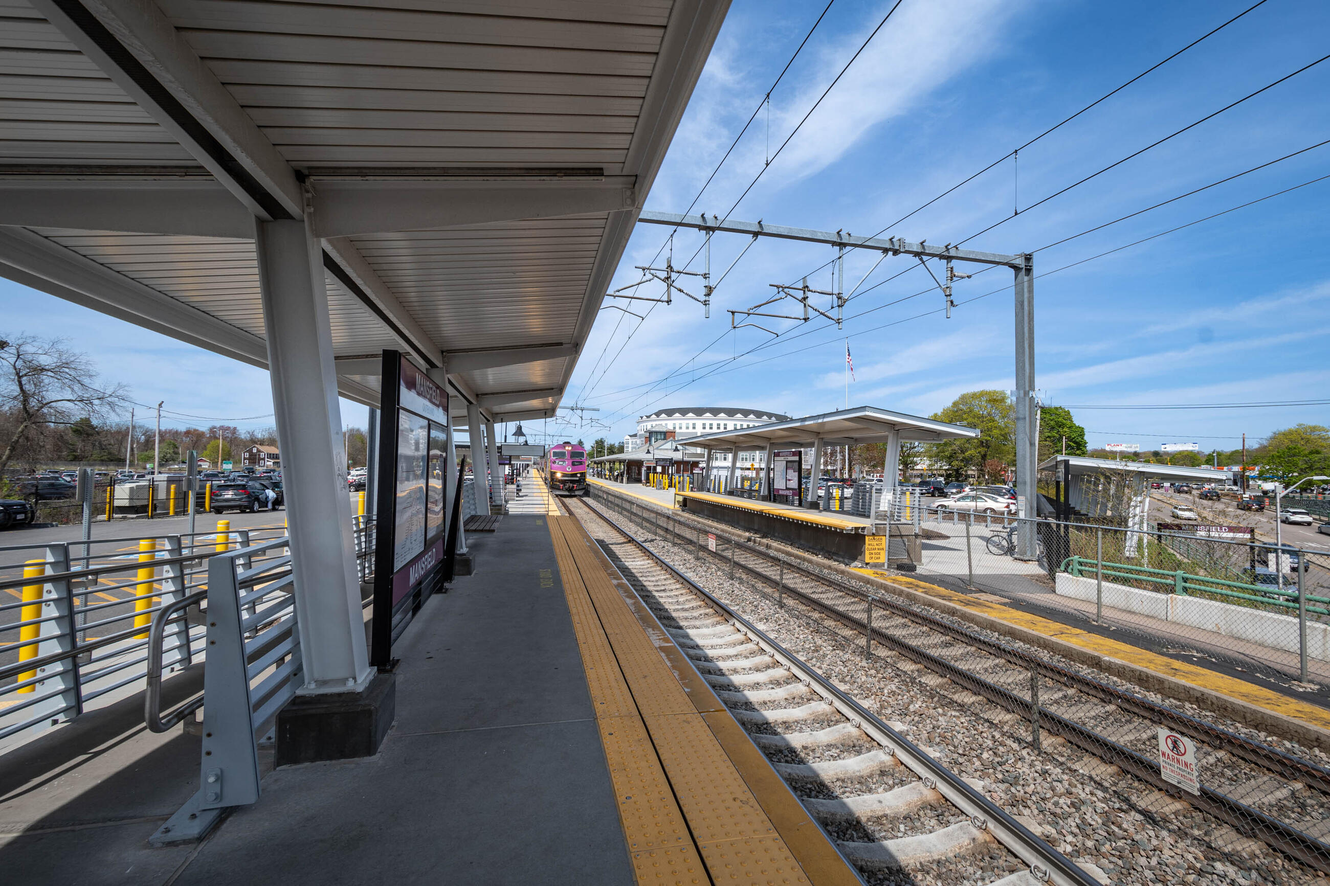A wide view of Mansfield station shortly after its accessibility upgrades were completed. The photo shows a long view of the outbound platform at left, with a train approaching. The track runs from the center to the lower right corner. The inbound platform is shown at right. 