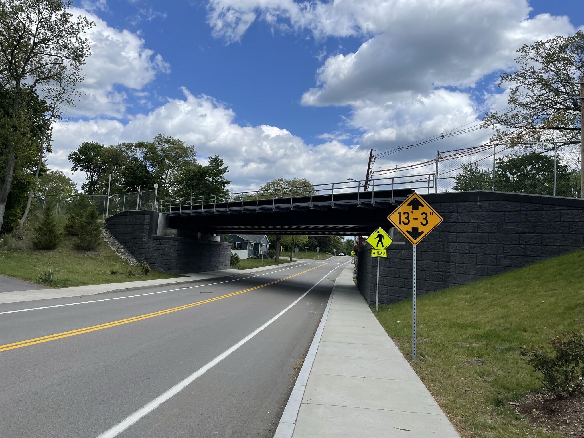 The completed bridge is viewed from a sidewalk down the road from the bridge. A sign showing that the bridge has a clearance of 13 feet and 3 inches is posted on the sidewalk just before the bridge; the previous bridge was under 11 feet high. 
