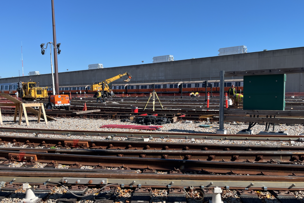 a yard of tracks on a cloudless day, orange line cars parked on the tracks in the furthest back, construction equipment spread out on various tracks