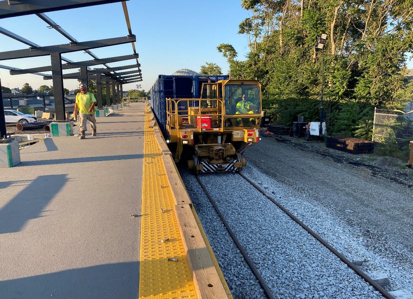 A construction worker drives a ballast spreader over the tracks at Fall River Station. Ballast are small rocks that serve as a bed for rail tracks and provide drainage and strength for heavy loads carried by trains. A second construction worker walks on the station platform alongside the ballast spreader.