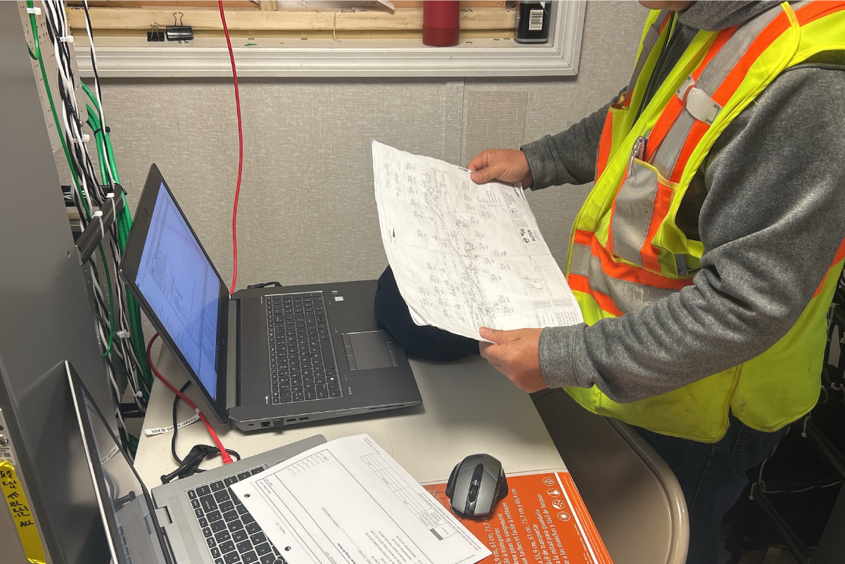 worker in reflective vest standing in front of two laptops looking at a sheet with a large diagram on it