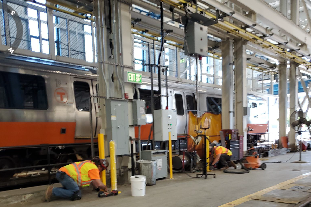 two crew members working on floor inside with orange line train in the background