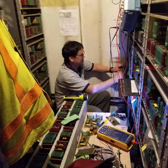 Inside the JFK/UMass signal house, a technician crouches among the signals to make repairs, with a safety vest and other equipment in the foreground